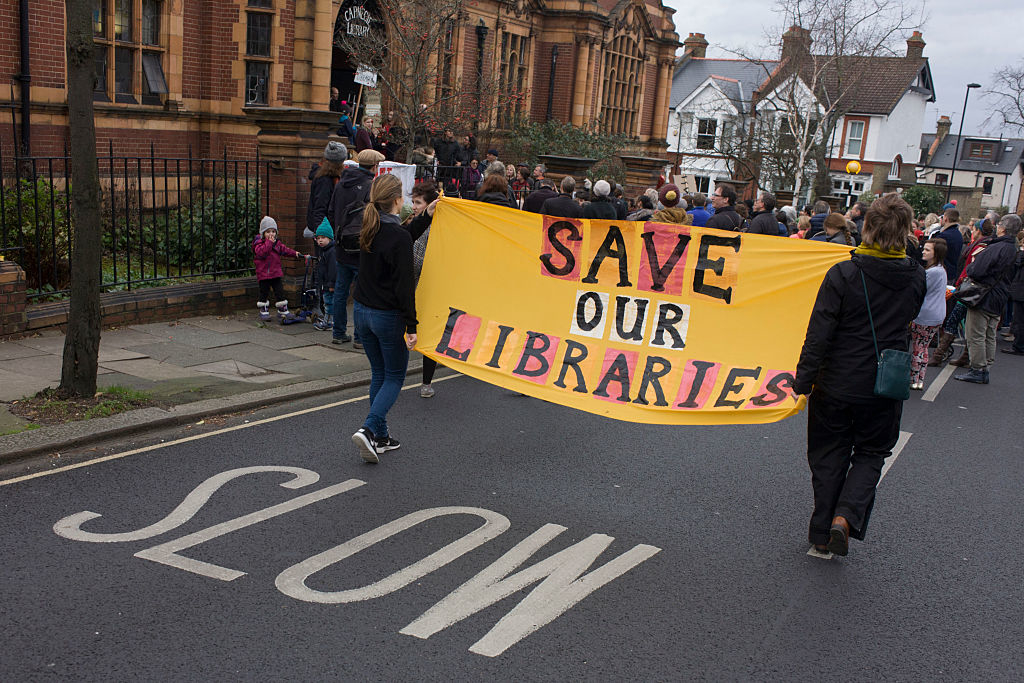 A crowd of people march down a street, holding a large yellow banner that reads &quot;Save Our Libraries.&quot;