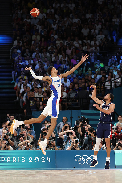 Victor Wembanyama of France (left) leaps to catch a basketball while Tyrese Haliburton of Team USA (right) shoots during the 2024 Paris Olympics in a packed arena