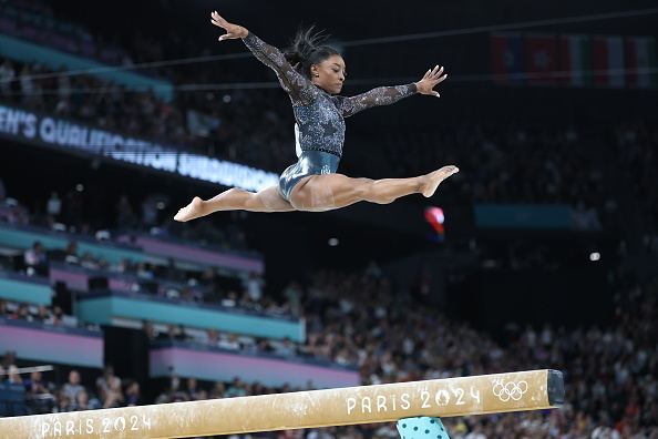 Simone Biles performs a split leap on the balance beam at the Paris 2024 Olympics during a gymnastics competition while a crowd watches