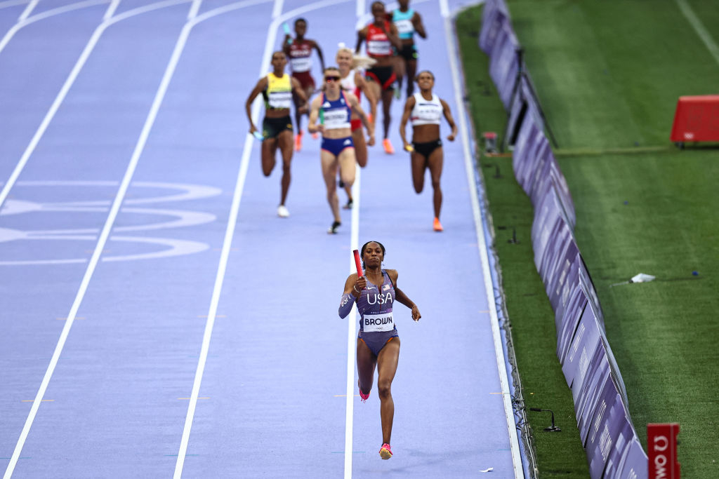 Athletes in a relay race on a track; a runner from Team USA is in the lead, holding a baton, followed by other competitors