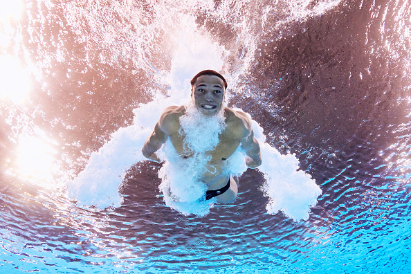 A diver is captured underwater during a dive, surrounded by a burst of bubbles, looking upwards towards the surface