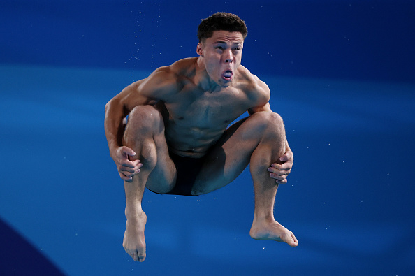 A diver mid-air with legs pulled to chest, making a concentrated facial expression, in a competitive diving event. The background shows the pool area