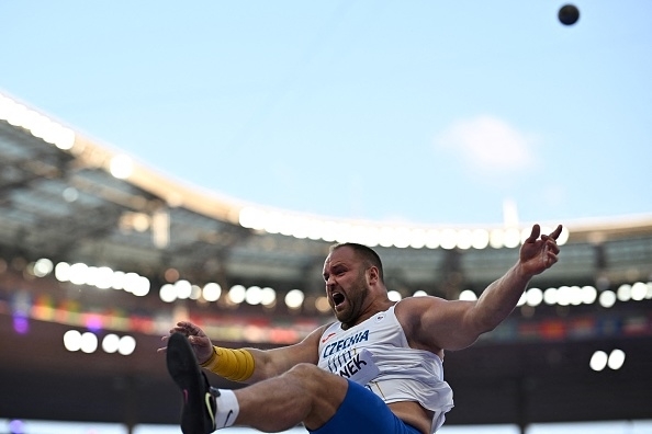 A male athlete mid-jump during a stadium event, wearing an athletic uniform with "Czechia" and "Stanek" visible, while a shot put flies in the distance