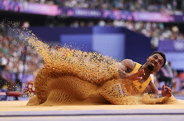 A sprinter is captured mid-jump during a long jump event at an outdoor sports competition, with sand flying around them upon impact