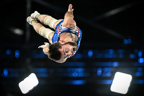 A gymnast performs a mid-air flip during a trampoline event, wearing a sleeveless bodysuit with a dynamic design. The image captures gymnastic precision and athleticism