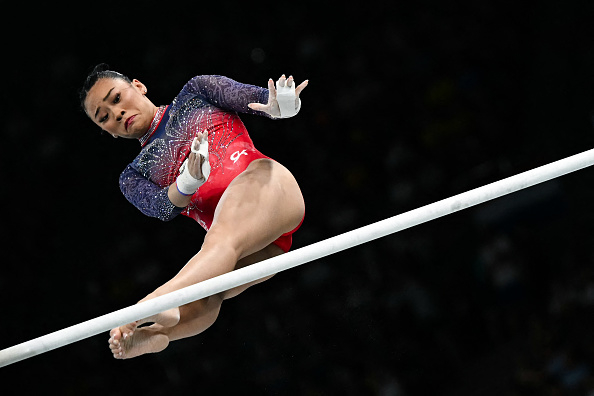 Suni Lee performs a difficult routine on the uneven bars during a gymnastics competition. She is in mid-air, showing her athletic prowess