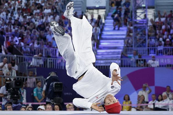 An athlete in white sportswear and a red beanie performs an impressive mid-air flip in a crowded arena