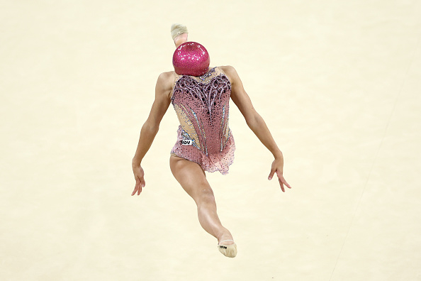 A gymnast performs with a ball balanced on their head, wearing a detailed costume during a rhythmic gymnastics routine