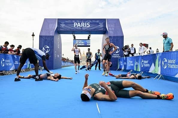Participants rest on the ground after completing a triathlon race under a Paris banner. Medics assist some athletes while others stand in the finish area