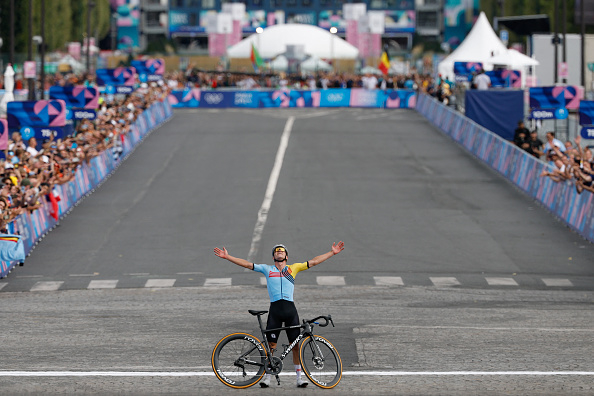 A cyclist stands triumphantly with arms raised, bike in front, on a race finish line, surrounded by cheering spectators. Bicycle race event