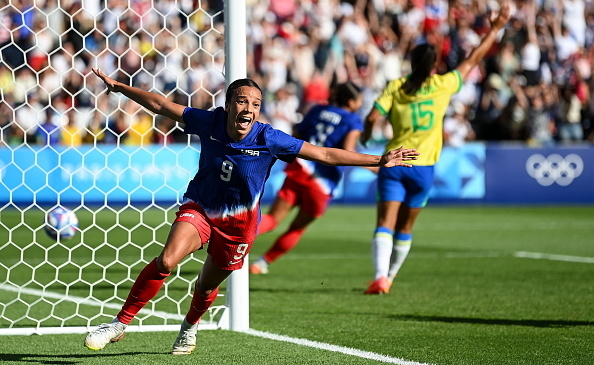 A soccer player, visibly excited, runs near the goalpost with outstretched arms during an Olympic match. Another player in background is seen celebrating