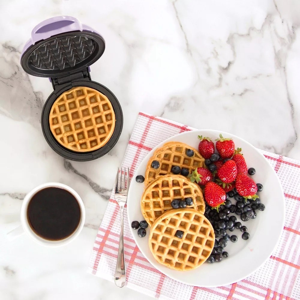 Small waffle maker with a cooked waffle, a plate with waffles, strawberries, blueberries, and a cup of coffee on a marble surface with a checkered napkin