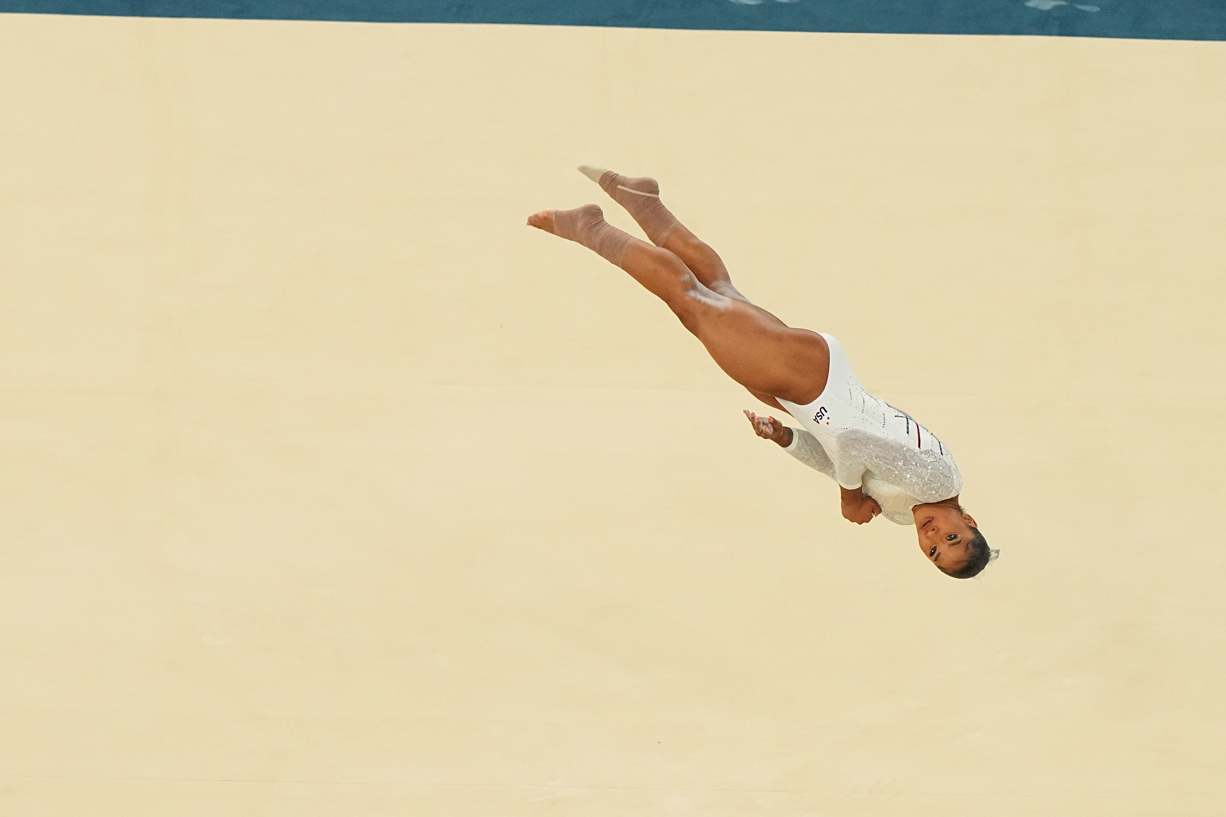 Jordan Chiles performs a mid-air flip during a gymnastics routine. She wears a white leotard decorated with sparkles