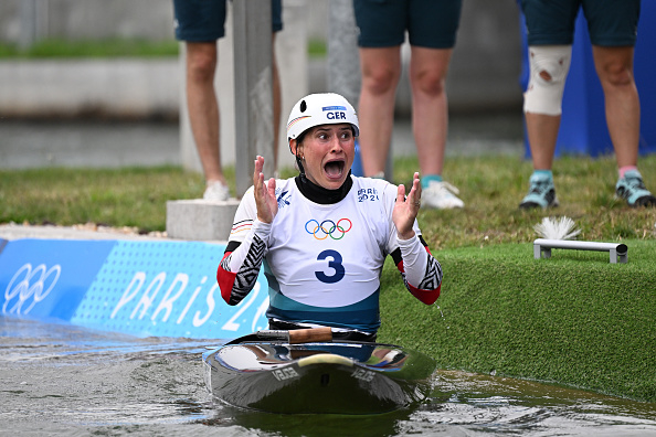 A female German athlete in a kayaking outfit, wearing a helmet and numbered bib, celebrates with surprise on her face after completing a race at Paris 2024