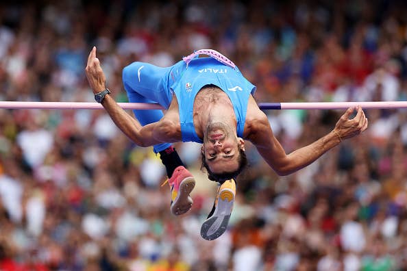 Gianmarco Tamberi executes a high jump at a sports event, wearing an Italy uniform. The crowd watches in the background