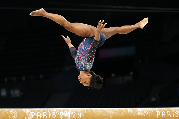 Simone Biles performs a mid-air flip above a balance beam, with "Paris 2024" visible on the beam