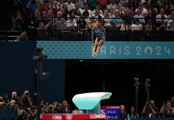 Simone Biles performs a mid-air flip during the vault event at the Paris 2024 gymnastics competition, with a large crowd watching in the background