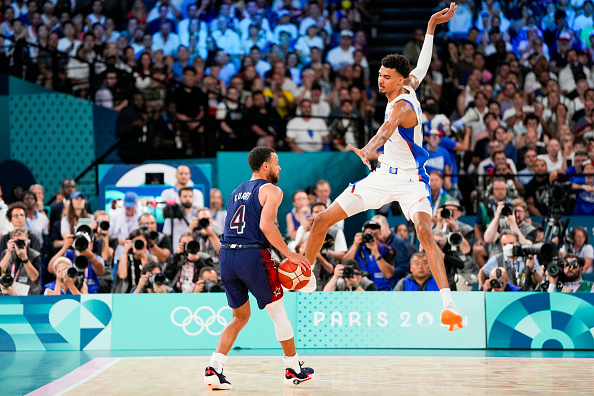 Victor Wembanyama and Rudy Gobert playing basketball at the Paris 2024 Olympics. Wembanyama is mid-jump on defense, while Gobert is dribbling the ball
