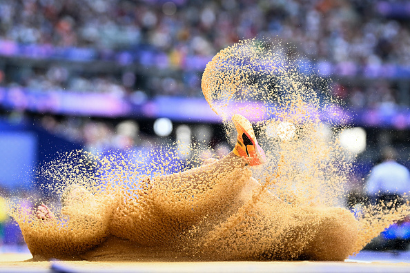 A track and field athlete lands in a sandpit during a long jump event, causing sand to fly up dramatically, with a crowded stadium in the background