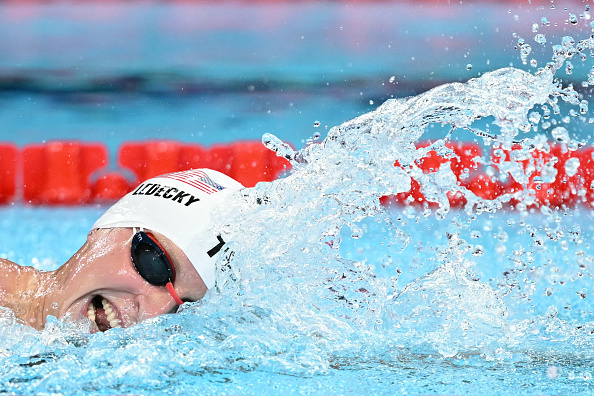 Katie Ledecky swimming in a competitive race, wearing a swim cap and goggles, with water splashing around her