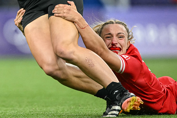 Rugby player Alisha Butchers in red kit held onto the leg of an opposing player during a tackle in a match, showing intense emotion