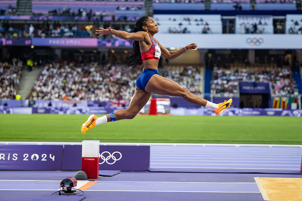 Olympic athlete in action, mid-air during a long jump attempt at Paris 2024 event, with stadium and spectators in the background