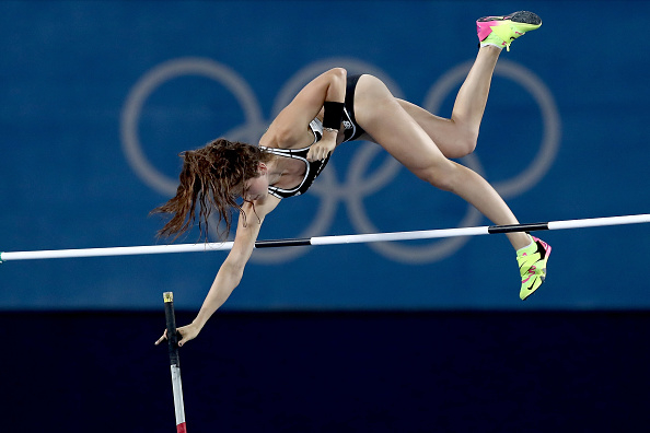 An athlete in mid-air while pole vaulting during a competition, reaching to clear the bar. The Olympic rings are visible in the background