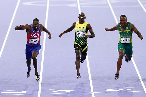 Noah Lyles, Julian Forte, and Akani Simbine sprinting during a track race. Lyles wears a USA uniform, Forte wears a Jamaica uniform, and Simbine wears a South Africa uniform