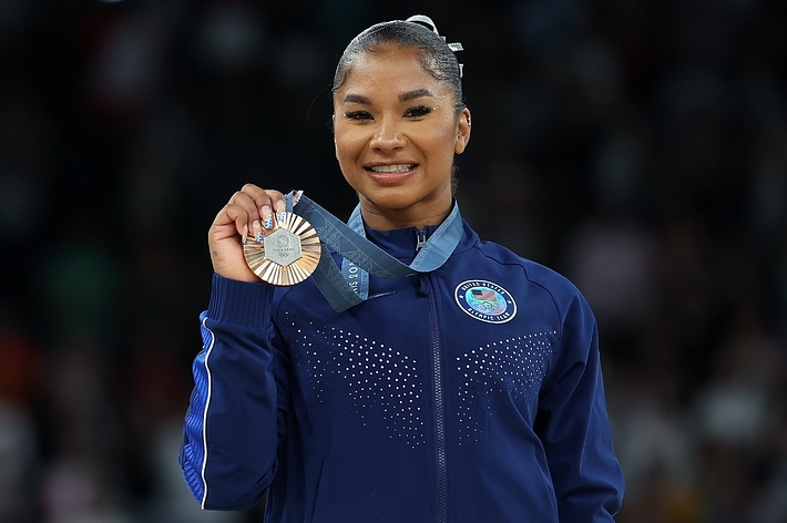 A woman, smiling and wearing a blue athletic jacket, holds up a silver medal