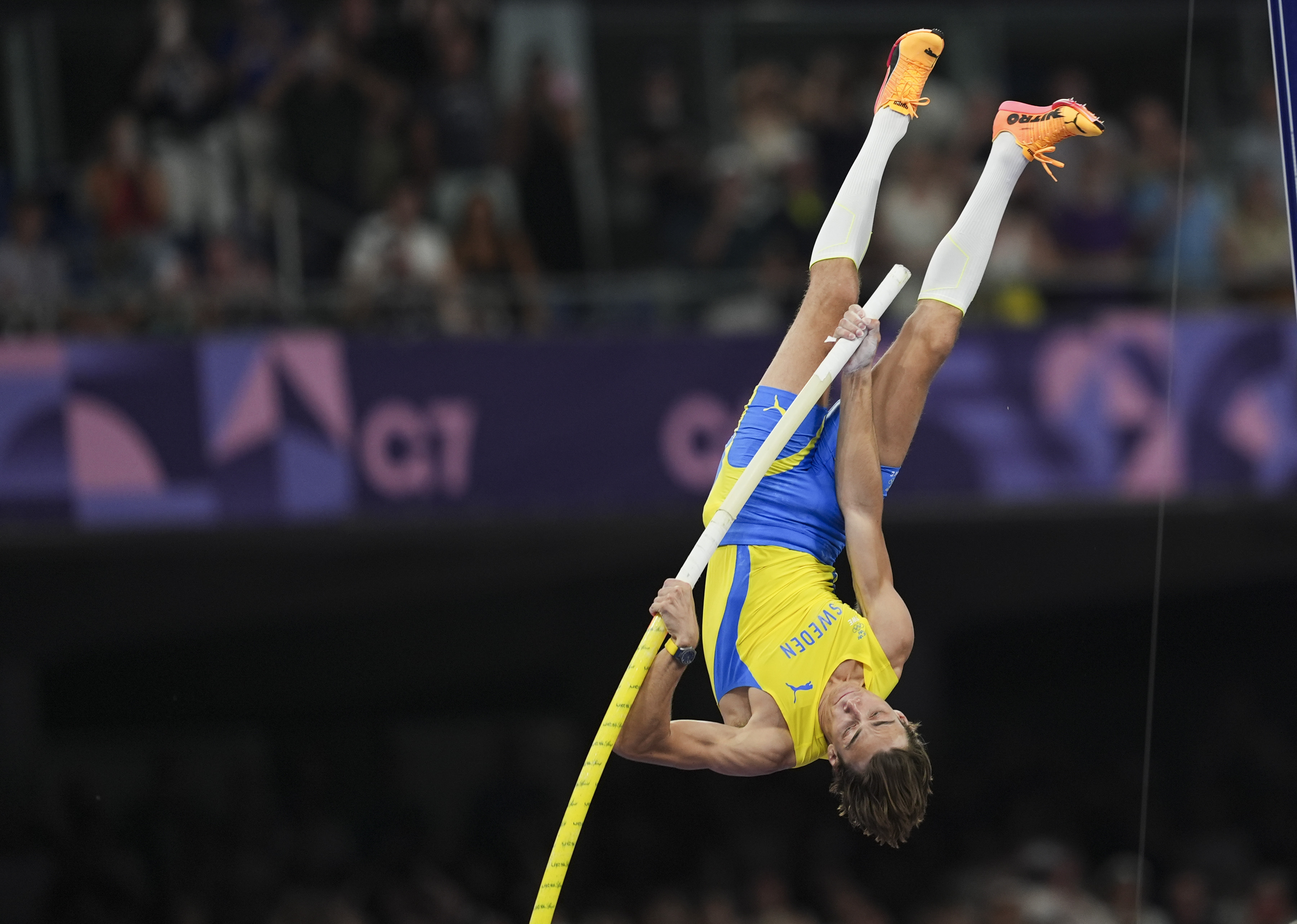 Pole vaulter Armand Duplantis in mid-air during a pole vaulting competition, wearing an athletic outfit and bright shoes