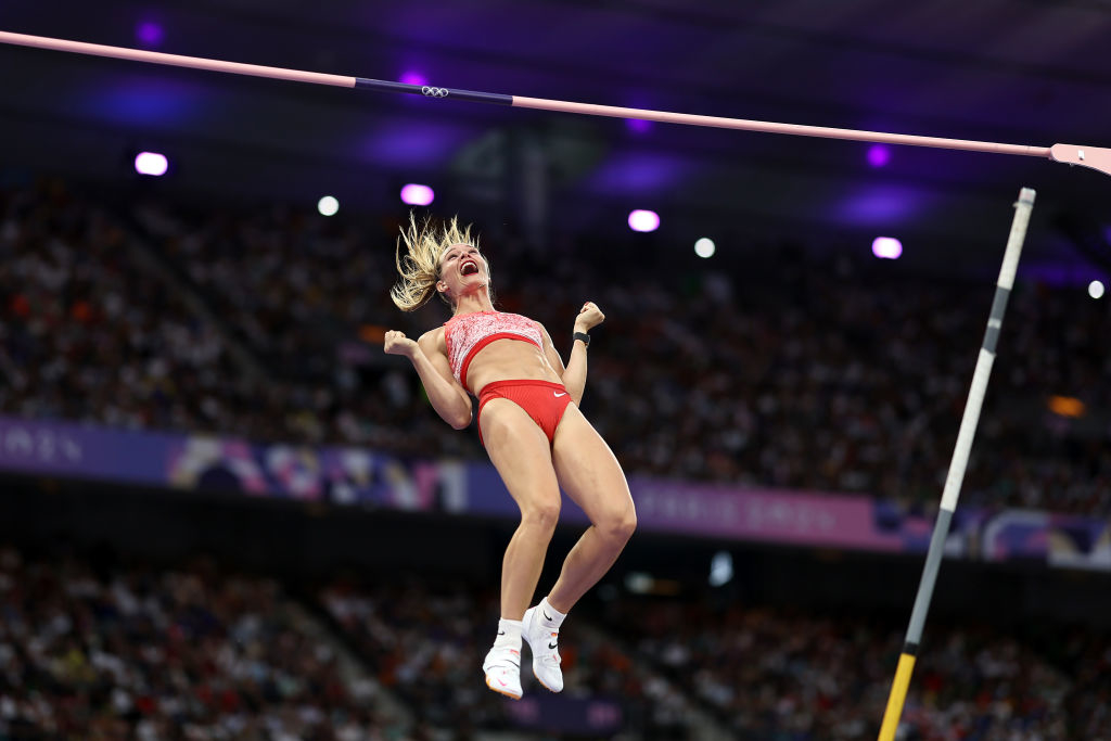 A female athlete in mid-air during a pole vault event, wearing athletic gear, appears excited. The stadium in the background is filled with spectators