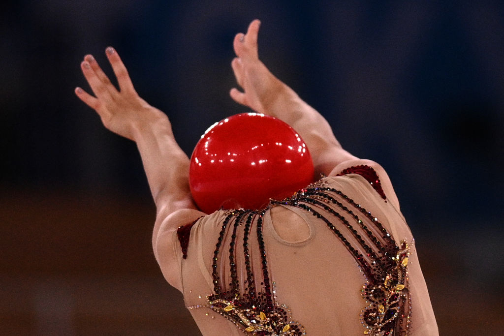 Person in an ornate costume holding a red ball above their head, arms extended, performing a rhythmic gymnastics routine