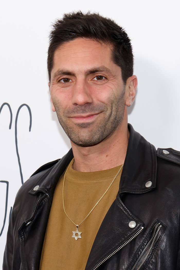 Nev Schulman wears a black leather jacket over a mustard shirt with a Star of David necklace, smiling at the camera in front of a white background