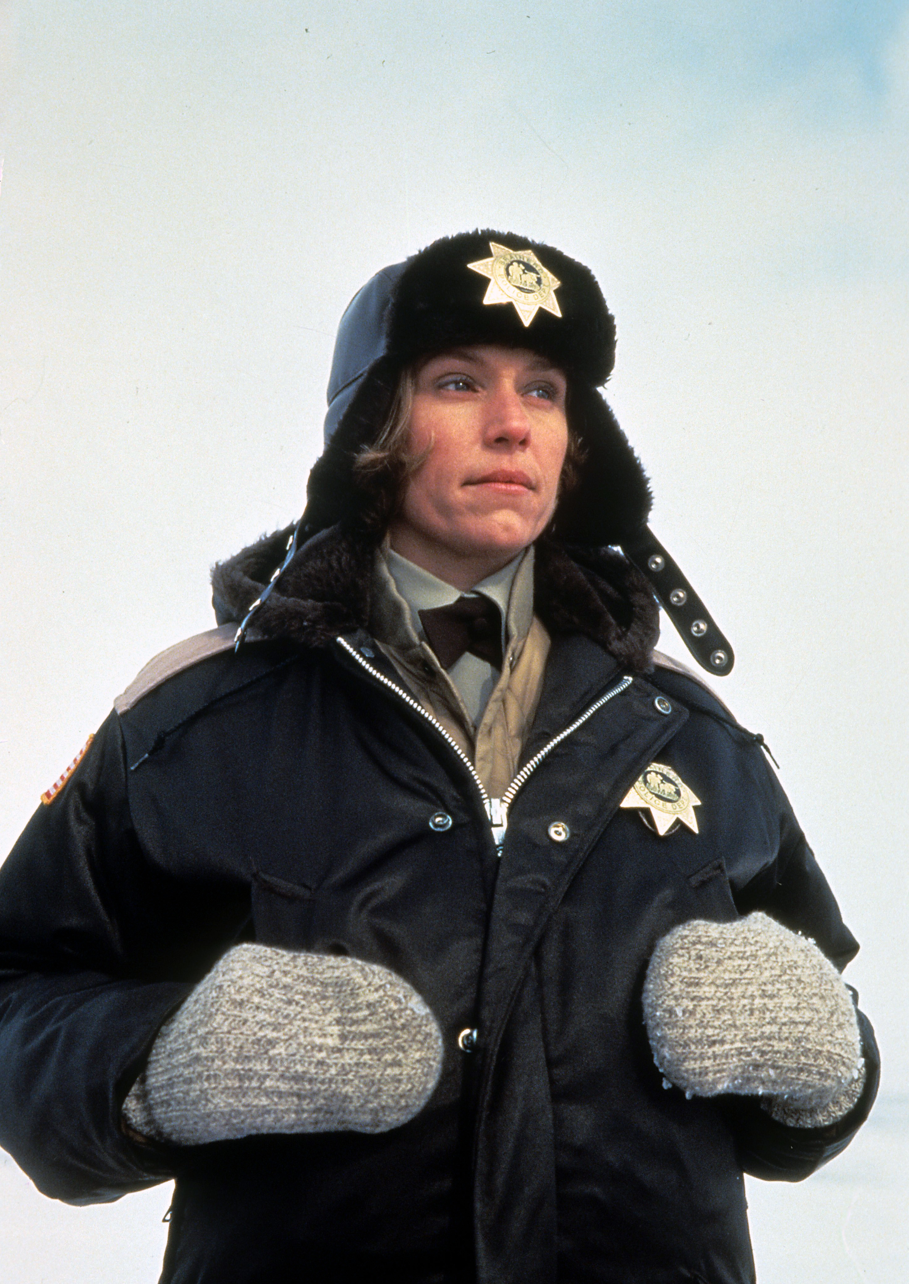 Frances McDormand dressed as a police officer, wearing a winter hat, a thick jacket, and mittens, as seen in her role from the movie "Fargo."
