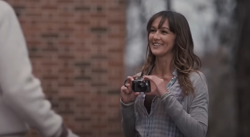 Sharni Vinson with long hair and bangs holds a camera in an outdoor setting with a brick wall in the background