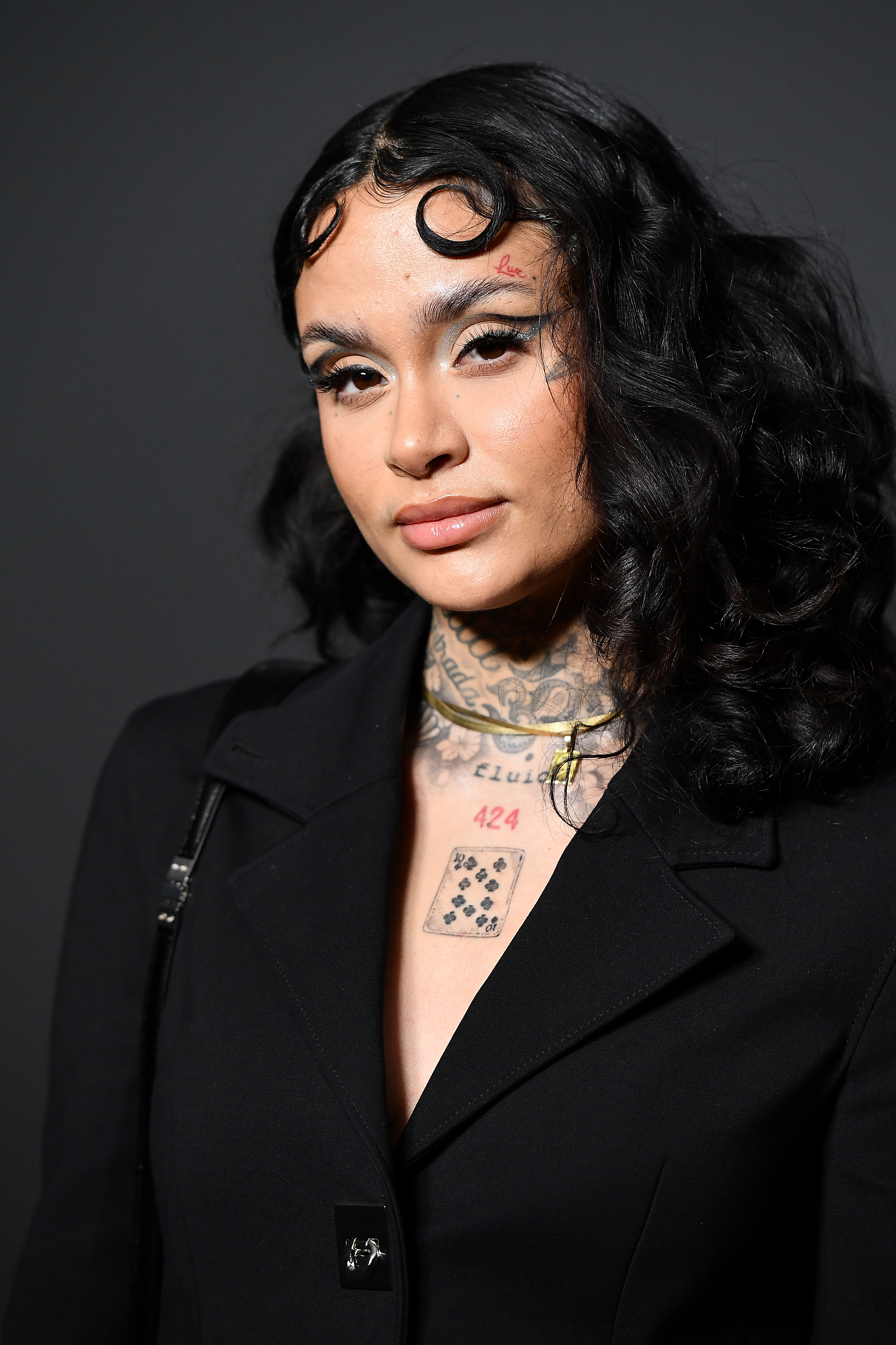 Kehlani poses against a simple background, wearing a black coat with unique tattoo designs on her neck and chest
