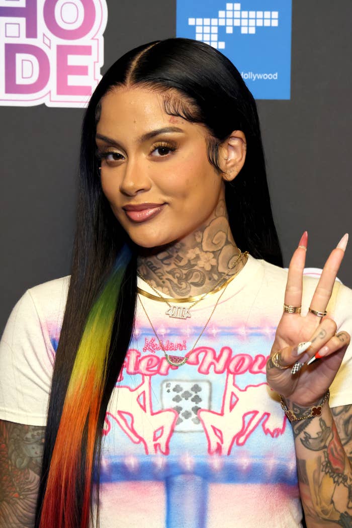 Kehlani, wearing a printed T-shirt with unicorns and a variety of jewelry, poses with a peace sign at a public event