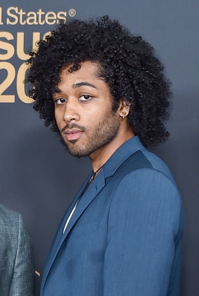 A person with curly hair poses in a blue suit in front of a backdrop for the United States Census 2020
