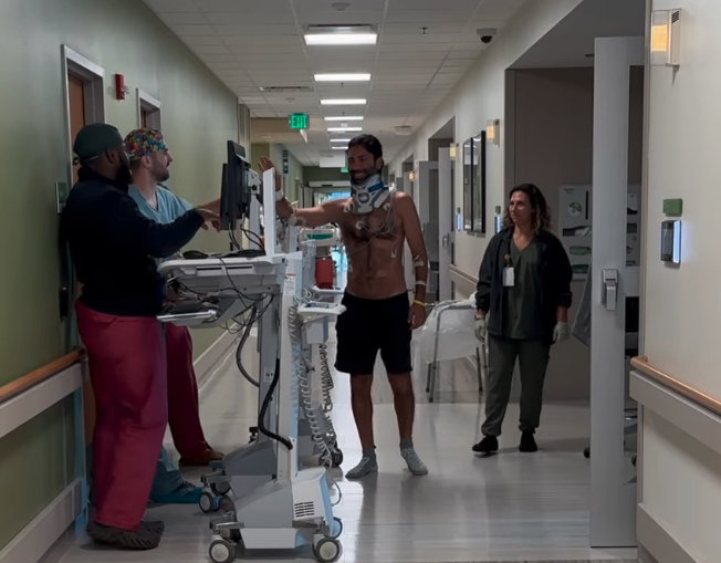 Keanu Reeves walks in a hospital hall, wearing a neck brace and connected to medical equipment, assisted by three medical staff members