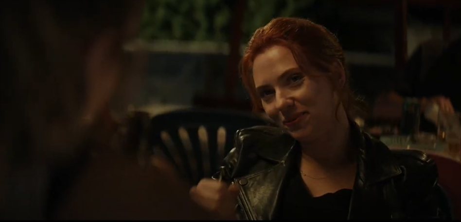 Scarlett Johansson in a black leather jacket, sitting and looking at someone off-camera in a casual setting with a subtle smile on her face