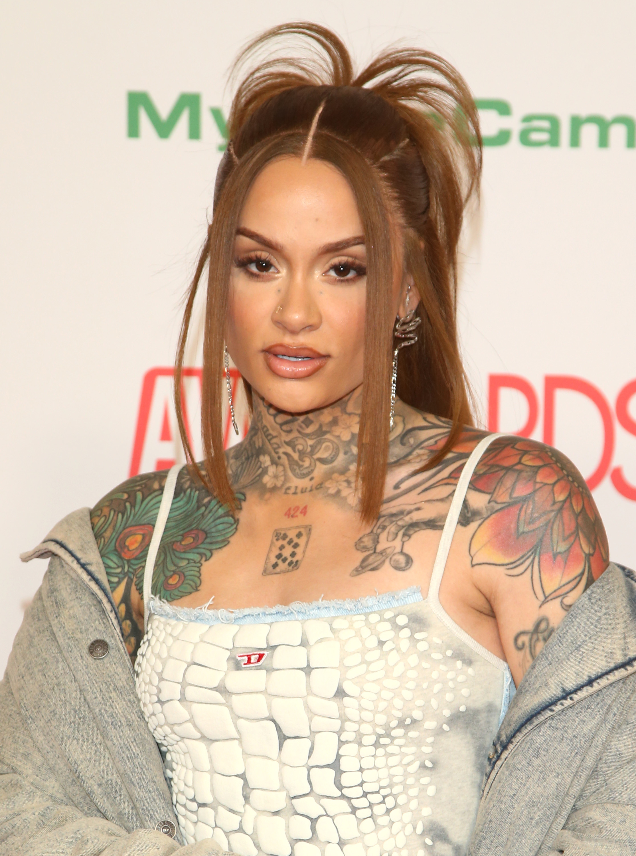 Kehlani at an awards show, wearing a white textured dress with spaghetti straps, denim jacket, and showcasing extensive chest and arm tattoos
