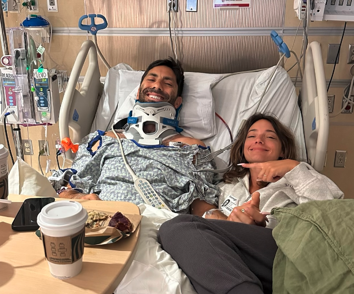 A man in a hospital bed wearing a neck brace, smiling, with a woman sitting beside him pointing at him. Coffee and food are on a table nearby