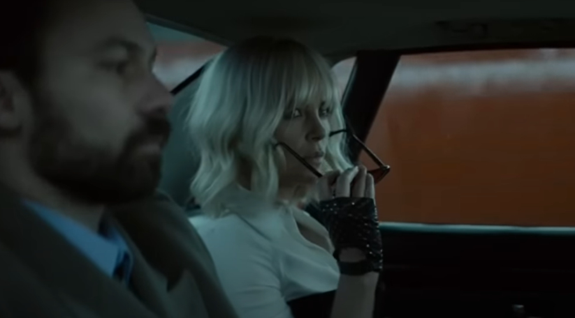 Charlize Theron sits in the backseat of a car, wearing sunglasses and a black-and-white outfit with studded black gloves. She looks to the side with a serious expression
