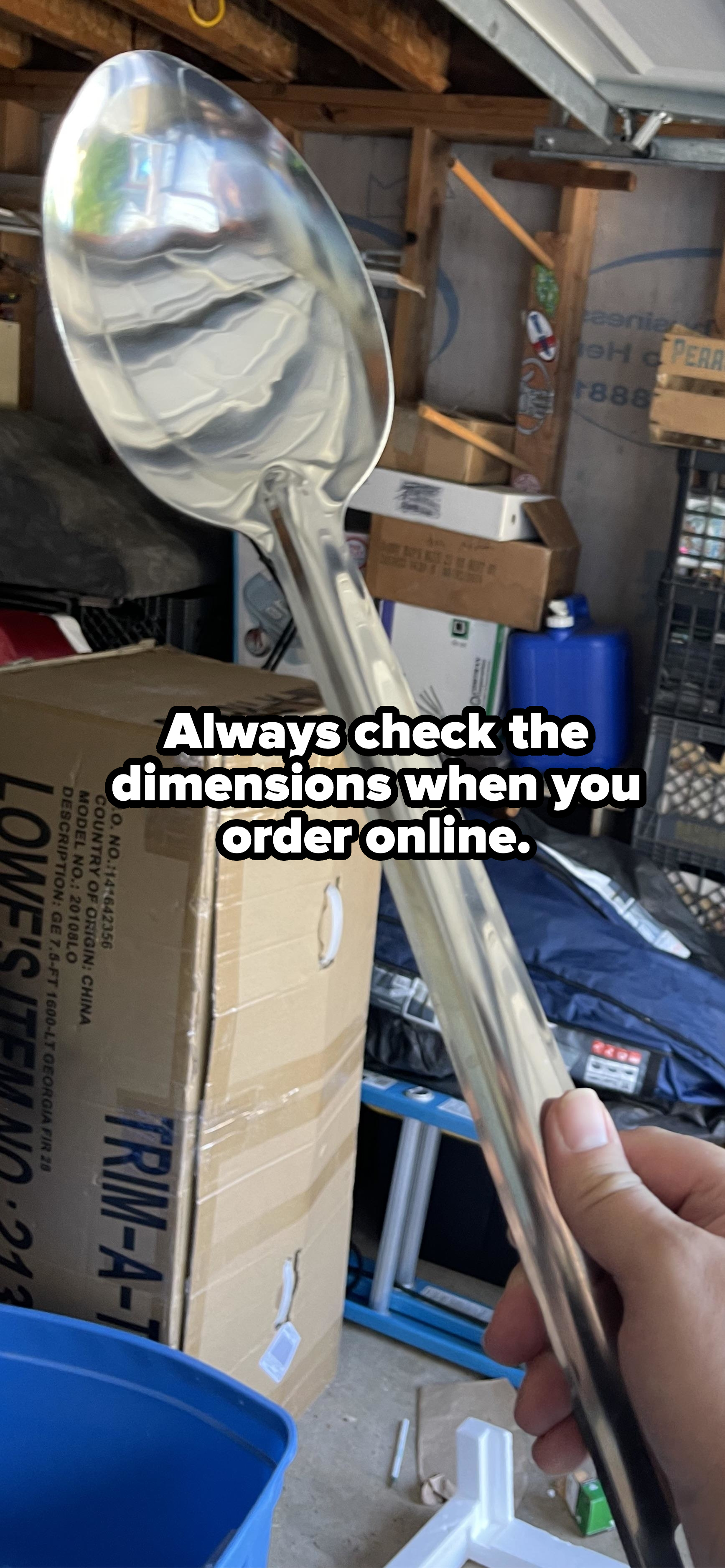 A hand holds a giant, reflective spoon in a cluttered garage. Various boxes and items are visible in the background