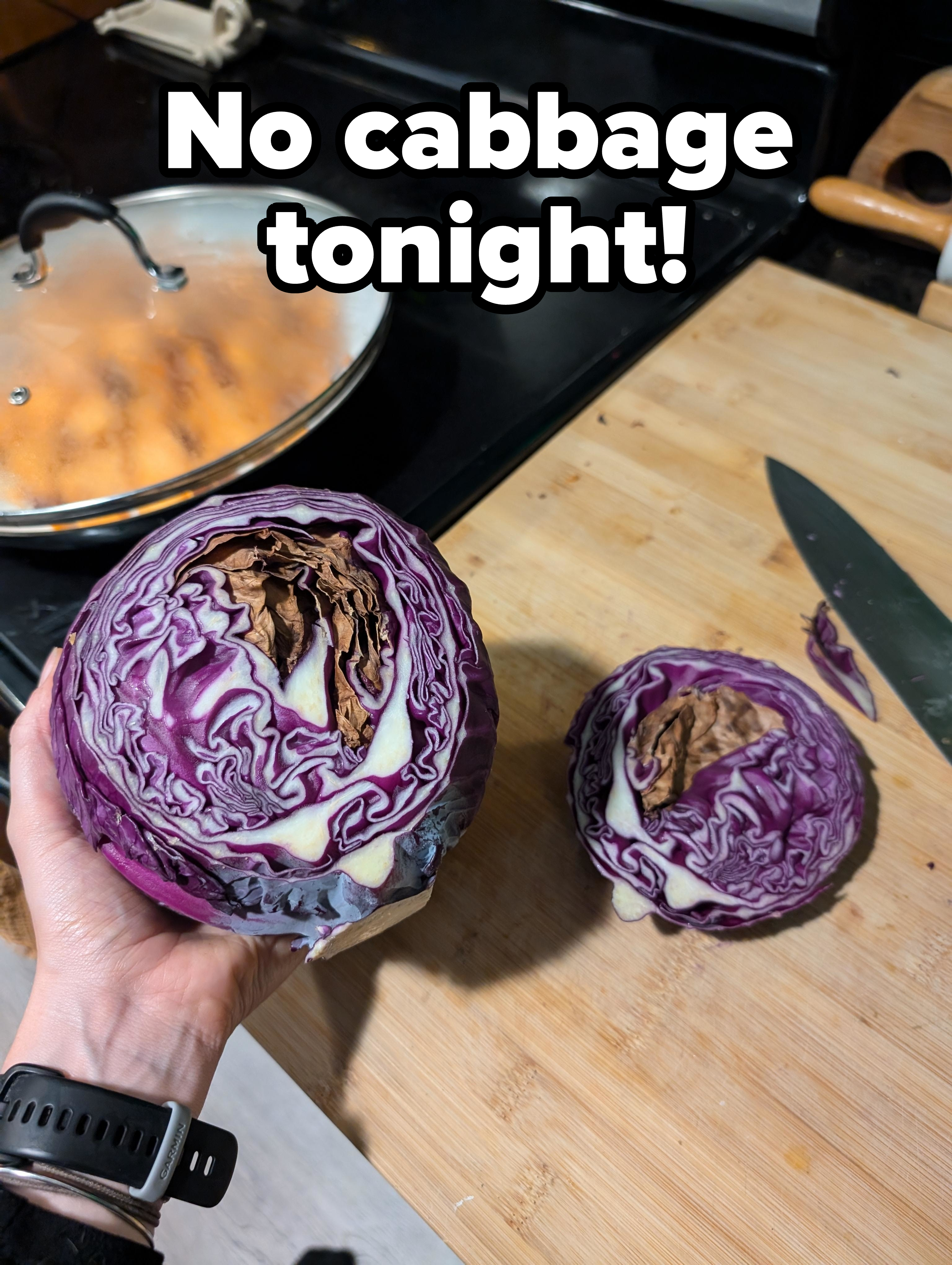 Person holding a halved cabbage over a cutting board, next to a knife and another halved cabbage on the board. Cooking pot and stove in the background