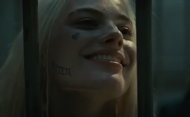 Margot Robbie in Suicide Squad with long, blonde hair, small heart tattoo on cheek, and "ROTTEN" tattoo on her jaw, smiles behind metal bars