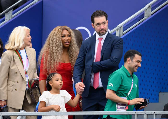 Serena Williams in a red dress, Alexis Ohanian in a suit, and their daughter Alexis Olympia Ohanian Jr. at an event. Another person stands nearby