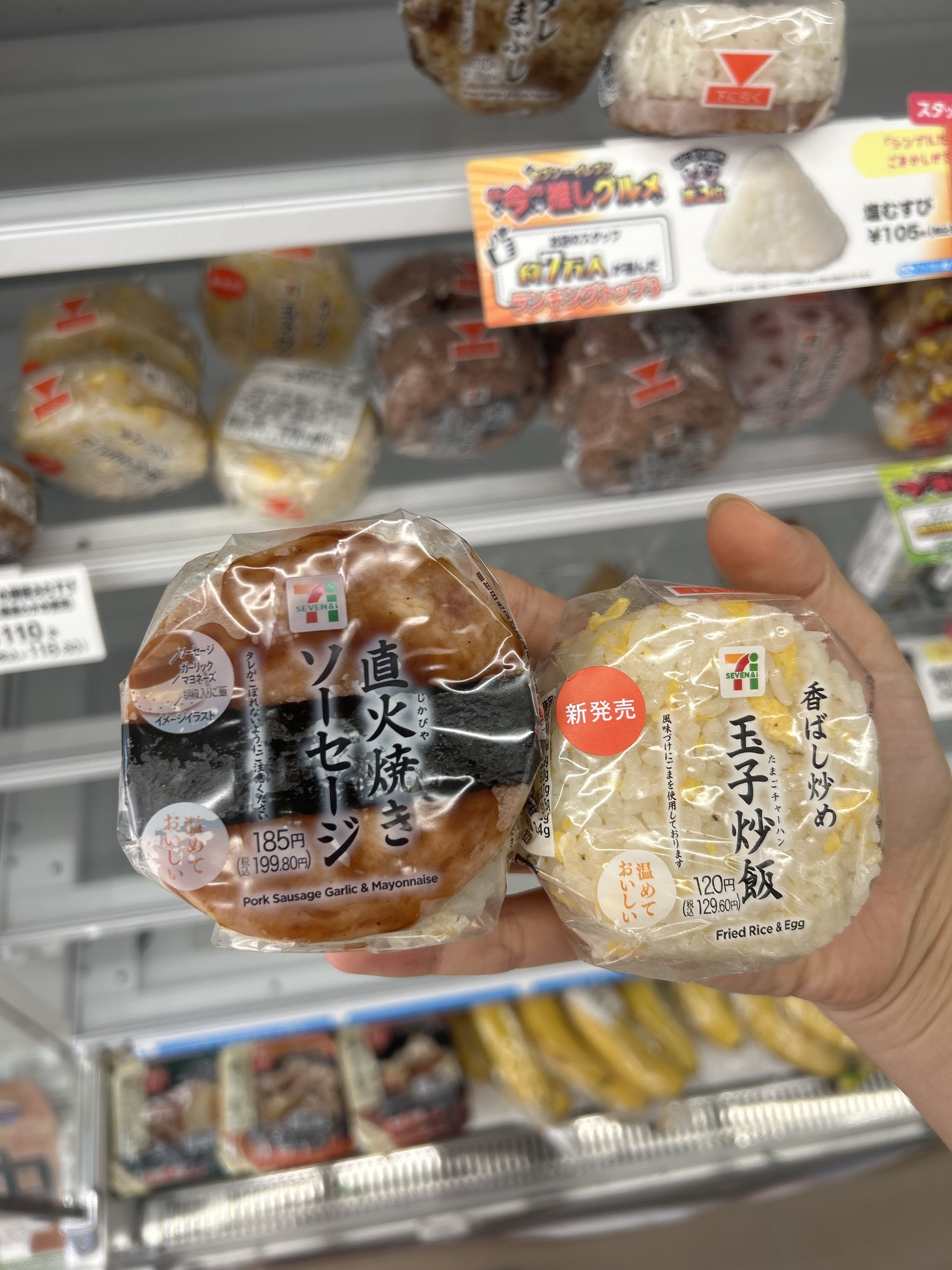 Two hands hold Japanese convenience store rice balls (onigiri) with labels: "Pork Sausage Garlic & Mayonnaise" on the left and "Fried Rice & Egg" on the right
