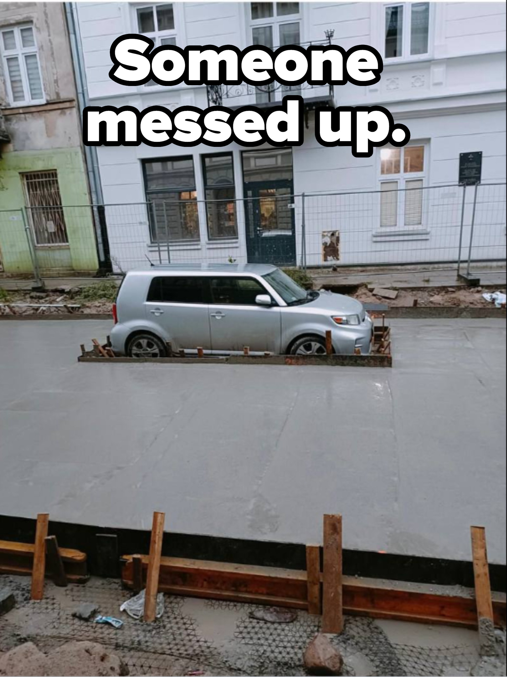 A silver car is stuck in the middle of a newly poured concrete street, in front of a white building with large windows and an ornate black balcony