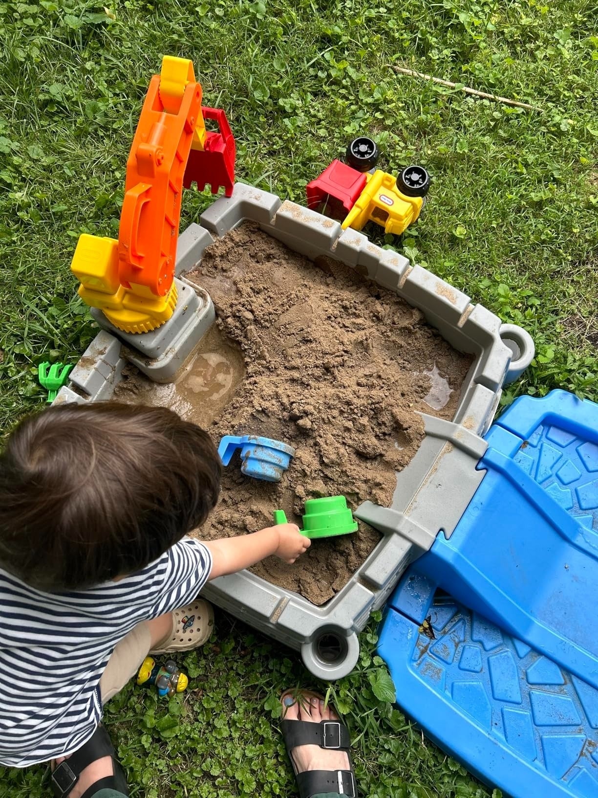 A kid  playing with a greenish  artifact  scoop successful  a sandbox. Various colorful toys, including a yellowish  crane and artifact  trucks, situation   the sandbox